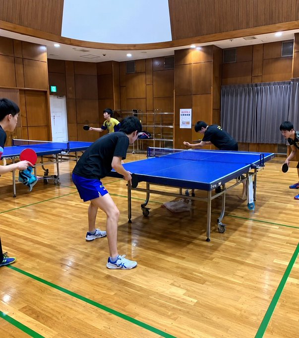 Several people playing table tennis in a wood-paneled gymnasium with multiple blue tables.