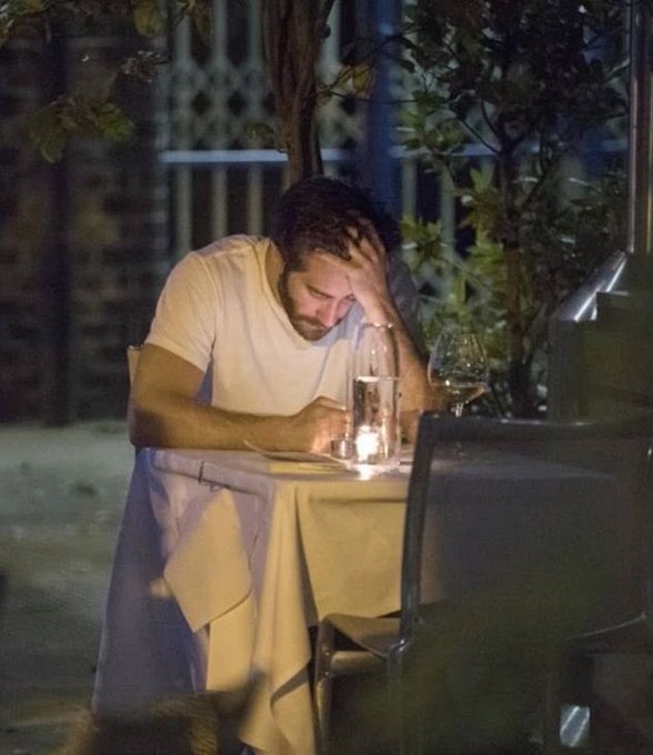 A man in a white t-shirt sits alone at an outdoor restaurant table at night, resting his head in his hand in a contemplative or dejected pose, with wine glasses and a candle light on the table.