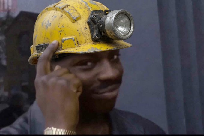 A man wearing a worn yellow mining helmet with a headlamp, giving a thumbs up gesture.