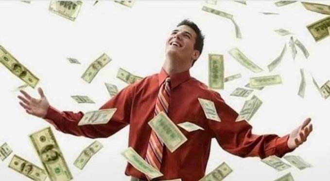 A smiling young man in a red shirt and striped tie stands with his arms outstretched as dollar bills rain down around him.
