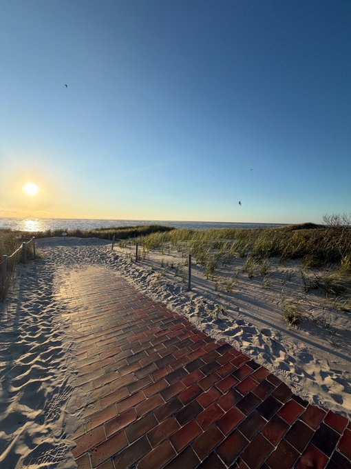 A scenic view of a brick pathway leading through sand dunes toward the ocean at sunset.