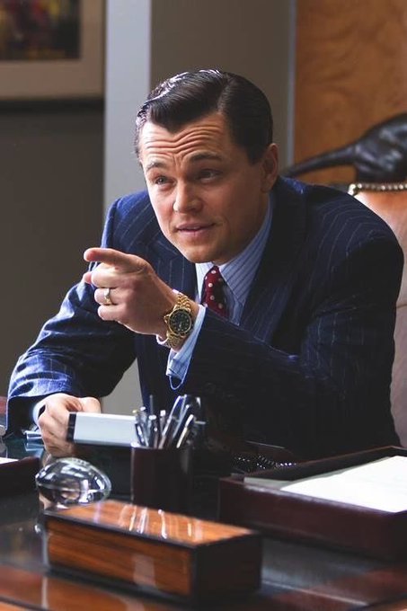 Leonardo DiCaprio in a dark pinstripe suit pointing his finger while seated at a desk with various office items.