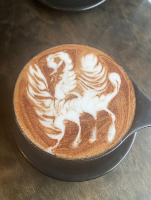 A cup of coffee with intricate latte art depicting a rooster or phoenix design, served in a black cup on a saucer.