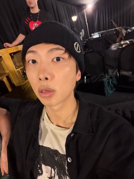 A young man wearing a black beanie and dark shirt takes a selfie indoors, with another person partially visible in the background near what appears to be musical equipment.