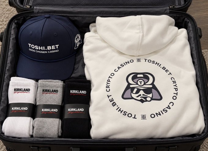 An open suitcase containing a navy 'TOSHI.BET Gods Chosen Casino' cap, three pairs of Kirkland Signature socks (white, gray, and black), and a white 'Crypto Casino' hoodie.