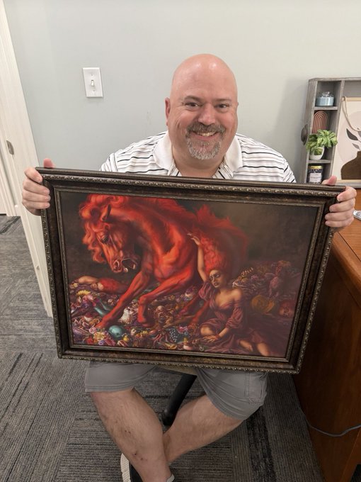 A bald man with a goatee, smiling while holding a framed painting depicting a dramatic red horse and figure with fruit.