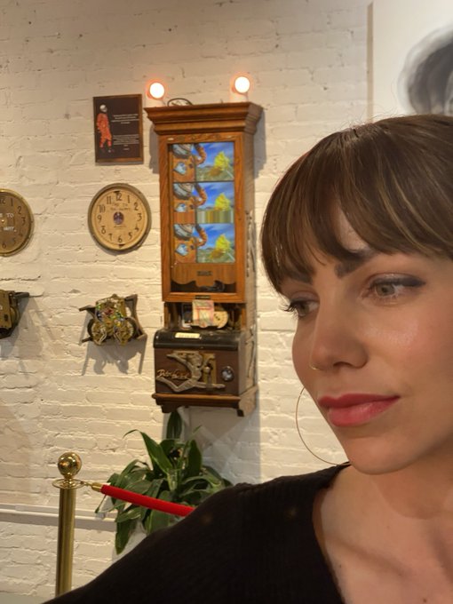 A woman with dark bangs and a nose ring takes a selfie in front of a white brick wall decorated with vintage clocks, a wooden 'Ripley's' game, and a red velvet rope.