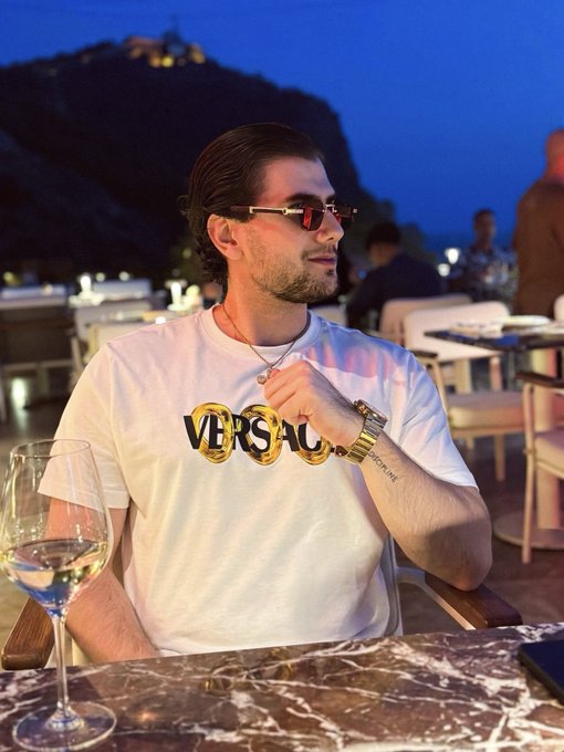 A man wearing a white "VERSACE" t-shirt and sunglasses sits at a marble table with a glass of white wine, posing for a relaxed evening photo with a lit hilltop in the background, fitting his casual "good morning" social media share.