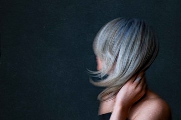 A side-profile image of a person with long platinum blonde hair, partially obscuring their face, against a dark background.