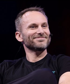 A man with short graying hair and a beard smiles subtly while wearing a black t-shirt, with a small microphone visible near his cheek. The image appears to relate to the post about Tether CEO Paolo Ardoino, suggesting this is Ardoino speaking at an event where he confirmed USDT coming to Bitcoin.