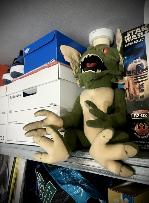 A Gremlin plush toy from Star Wars sitting on a shelf next to a 'STAR WARS ACTION COLLECT' R2-D2 box and various storage boxes.