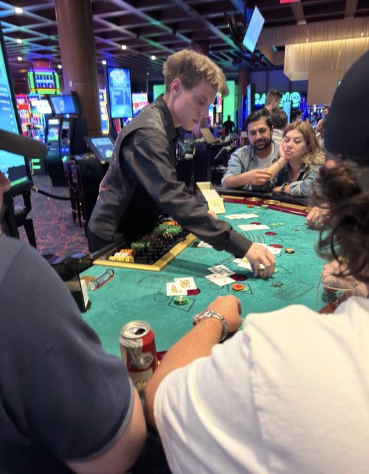 A casino scene where a young male dealer in a dark jacket is dealing cards at a green blackjack table while players watch, with a red Coca-Cola can on the table and slot machines visible in the background.