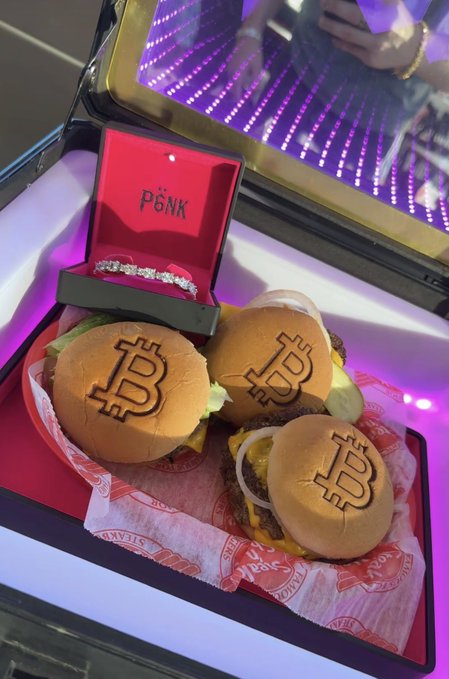 An open pink jewelry box holds a diamond bracelet alongside the word "PINK," resting in a tray with three small burgers branded with a "B" logo, all illuminated by purple LED lights.