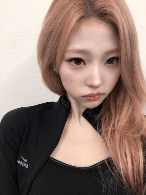 A close-up selfie of a young woman with long peach-pink hair, dramatic eye makeup, and a black athletic zip-up jacket with 'CALM' text visible on the sleeve.