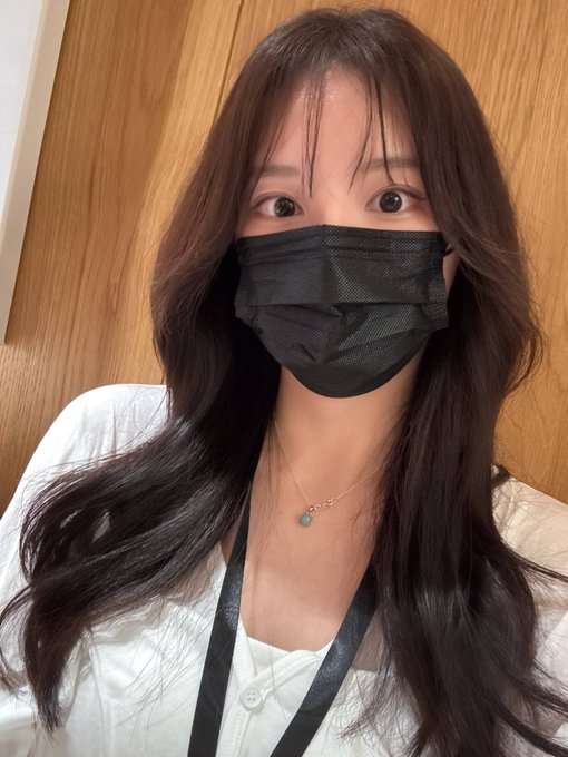 A young woman with long brown hair and bangs wearing a black face mask and white top takes a selfie, likely at a Token2049 event in Singapore where she's hoping to meet fellow attendees. Her friendly pose invites connection at the crypto conference.