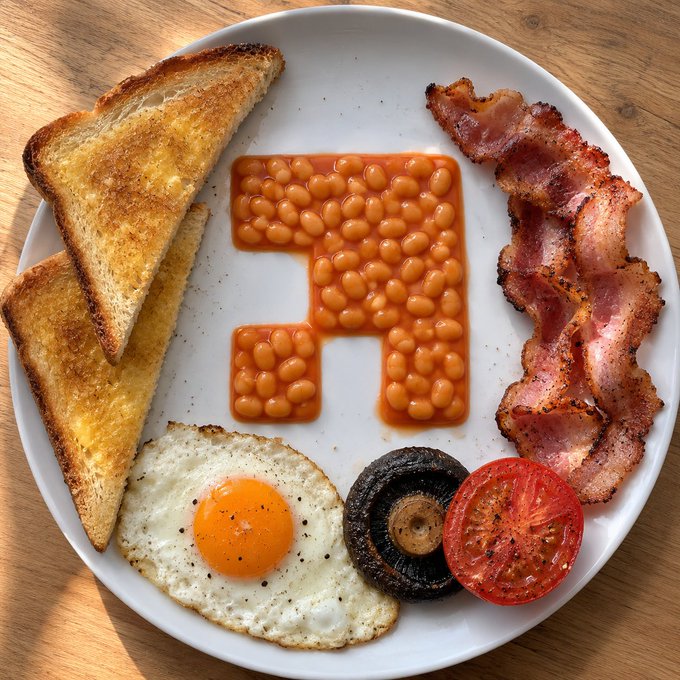 A full breakfast plate featuring toast, bacon, a fried egg, a grilled mushroom, and a grilled tomato, with baked beans artfully arranged into the shape of the number '1'.