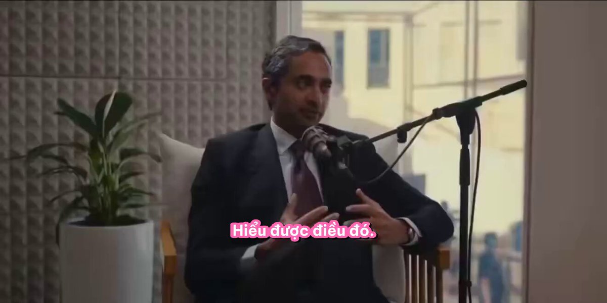 The image features a man sitting in an office chair, wearing a suit and tie, with his hands placed on his lap as he speaks into a microphone. The background of the image shows a window overlooking a city street, providing a view of buildings outside. There is also text visible in the image that reads "100K BITCOIN VẪN CHƠI VƠI 𝐁𝐓𝐂 파이썬 가능 경로 구분 걸리", which could be a description of the man's suit or an advertisement for a product.