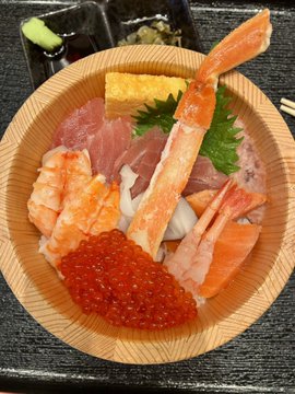 A Japanese seafood donburi bowl featuring crab legs, shrimp, raw tuna, tamagoyaki (egg), salmon roe, and garnished with parsley, served with a side of soy sauce and wasabi.
