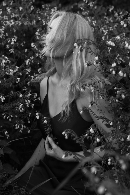 A black and white photo of a woman wearing a dress standing behind some flowers that have buds sprouting out from them with her hair blowing around. The photo has been edited, giving it an artistic feel, making it a striking piece of art.