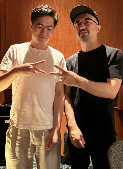 Two men, one in a white t-shirt and glasses and the other in a black cap and shirt, stand side by side both making finger gun gestures.