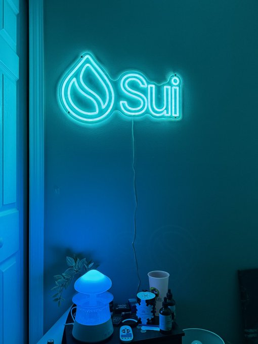 A blue neon sign of the Sui blockchain logo mounted on a dark wall, glowing above a table with various small items including bottles and figurines.