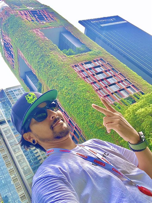 A man in a cap, sunglasses, and lanyard flashes a peace sign in front of the vertical greenery of Singapore's Oasia Hotel Downtown, alongside the "SHUI ON" skyscraper nearby, capturing a leisurely morning ("Gm") sightseeing moment in the city.