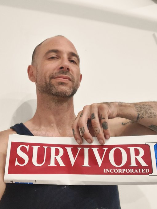 A man with a shaved head and tattoos holding up a red and white banner that reads 'SURVIVOR INCORPORATED' while looking at the camera.