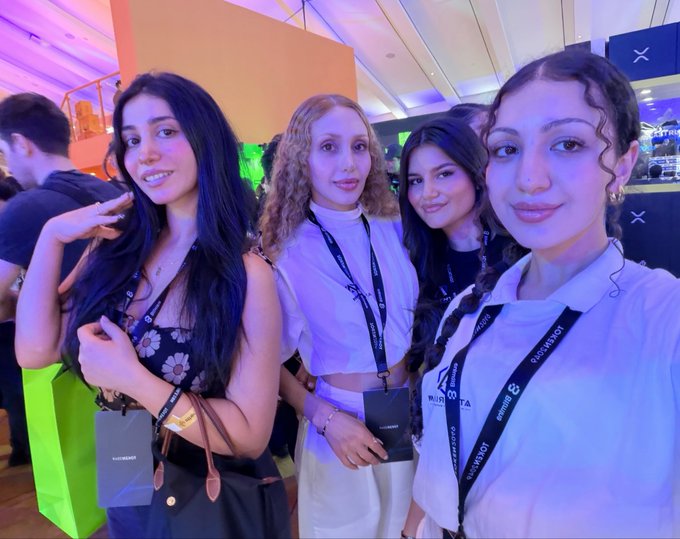 Four young women wearing 'TOKEN2049' lanyards smile together at a crypto conference event, with one holding a green gift bag.