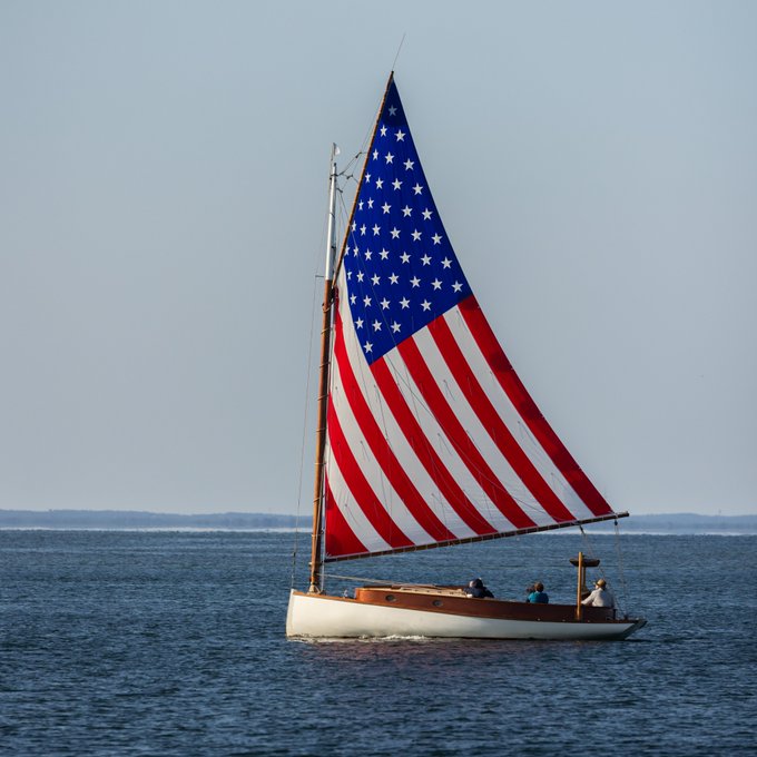 A sailboat with a large American flag-patterned sail carries several passengers across a calm body of water.