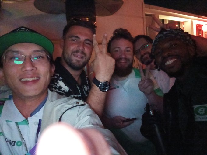 A group selfie of five smiling people at a social gathering, with several making peace signs and one wearing a cap and lanyard.