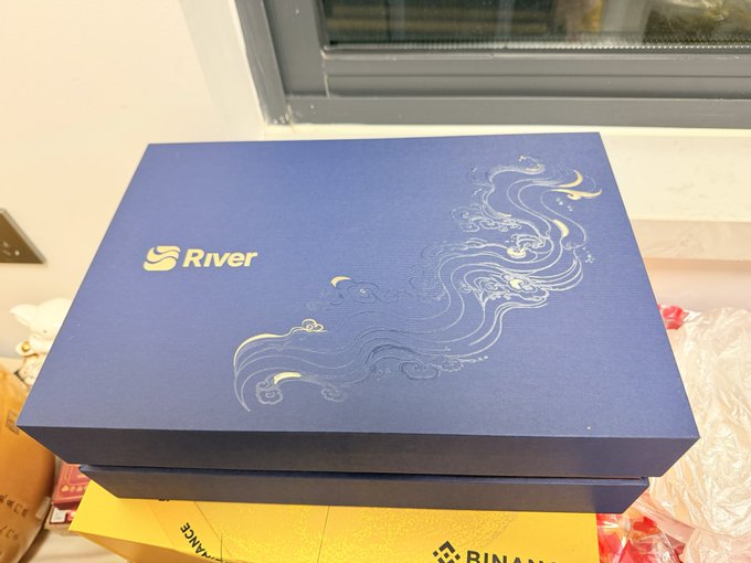 A blue "River" branded gift box with elegant cloud and moon designs sits on a yellow Binance package, matching the post about receiving a tea set gift from River and enjoying tea while watching $RIVER price charts during the National Day holiday.