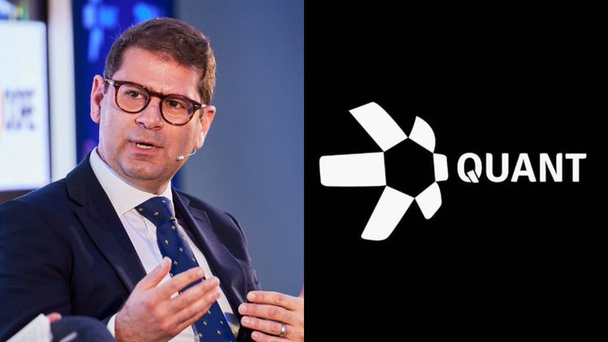 A man in glasses and a suit speaks on stage, paired with the white QUANT logo featuring a starburst symbol on a black background.