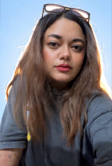 A close-up selfie of a woman with long wavy brown hair, sunglasses perched on her head, wearing a dark gray t-shirt against a clear blue sky background.