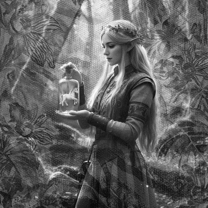 A black and white fantasy image of an elf-like woman with long blonde hair and a leaf crown, holding a glass bottle containing a small horse figurine, against a heavily patterned tapestry background.
