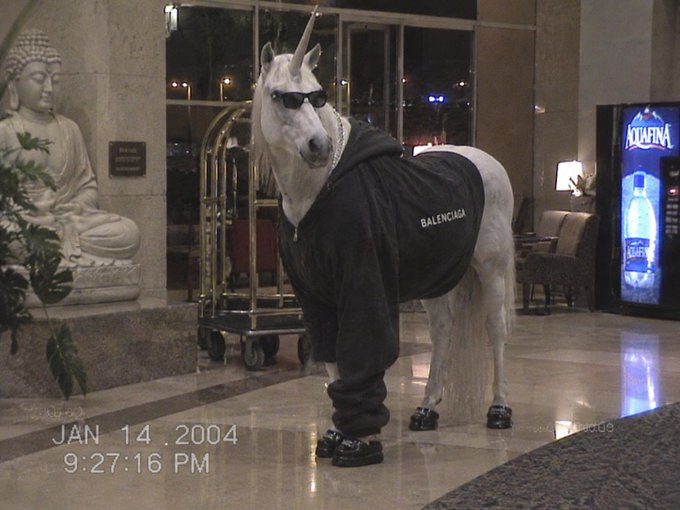 A white unicorn wearing sunglasses, a thick chain necklace, a black Balenciaga hoodie, and black sneakers stands in a hotel lobby near a Buddha statue and a luggage cart. The timestamp reads 'JAN 14, 2004 9:27:16 PM'.