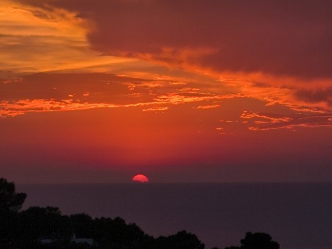 A scenic photograph of a red sun setting over the ocean, with dramatic orange and red clouds streaking across the sky above a dark silhouetted coastline.