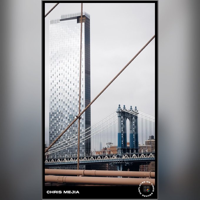A photograph by Chris Mejia taken on a Google Pixel, showing a modern glass skyscraper viewed from the Manhattan Bridge with the bridge's suspension cables and iconic blue towers visible, framed in black with a Shot on Google watermark.