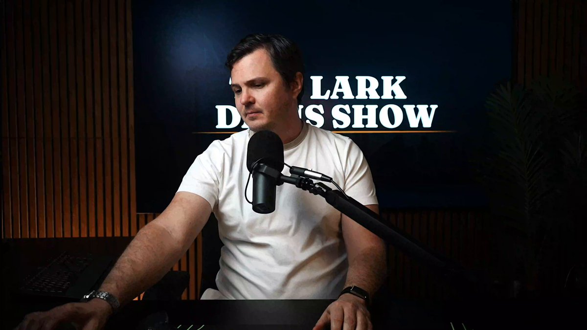 The image shows a man sitting at a desk with a microphone, likely preparing for or just finished recording a podcast episode related to cryptocurrency. The background features a television screen displaying the words "Lark Docks Show" along with some stock updates. The focus of the image is on the man and his equipment, emphasizing the importance of the content being discussed in the podcast.
