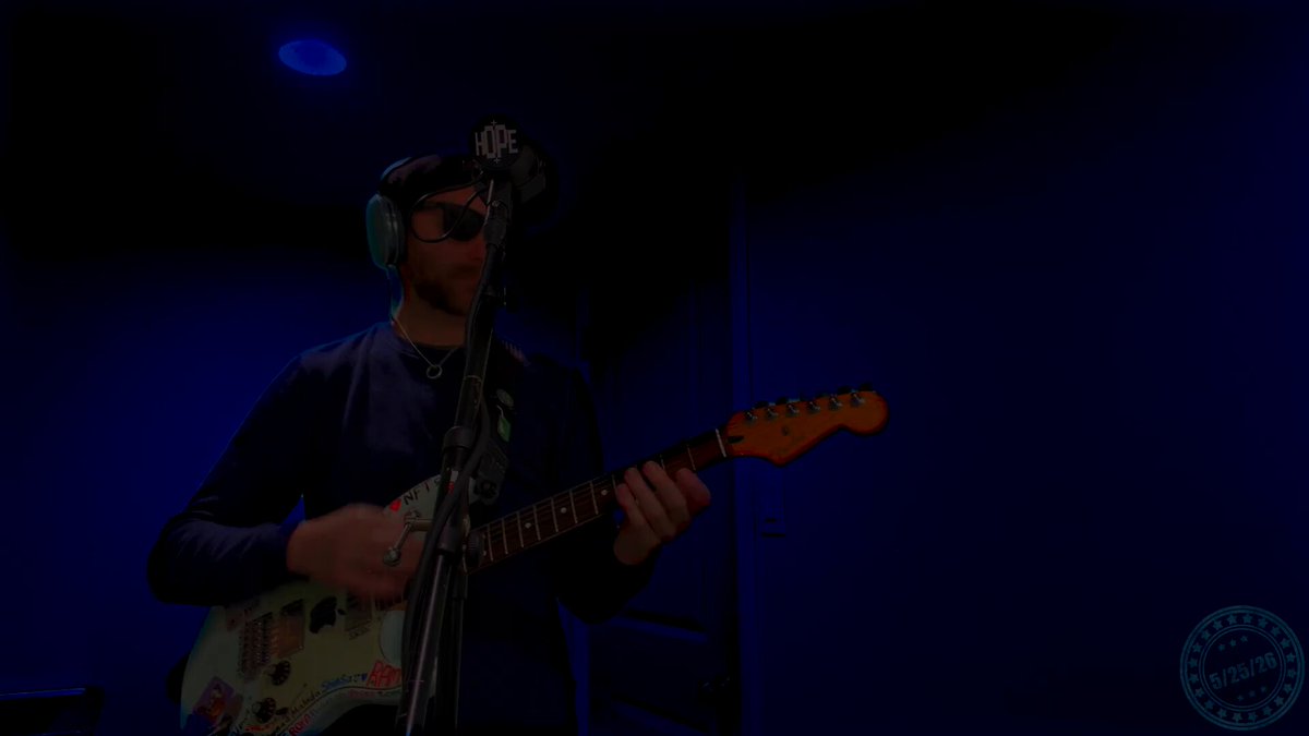 The image features a man playing an electric guitar, wearing headphones and standing next to a microphone. The scene takes place in a dark room with blue walls, creating a moody atmosphere. There are stickers on the guitar, adding a personal touch to the instrument. The man is holding the guitar while singing into it, showcasing his talent as a musician.