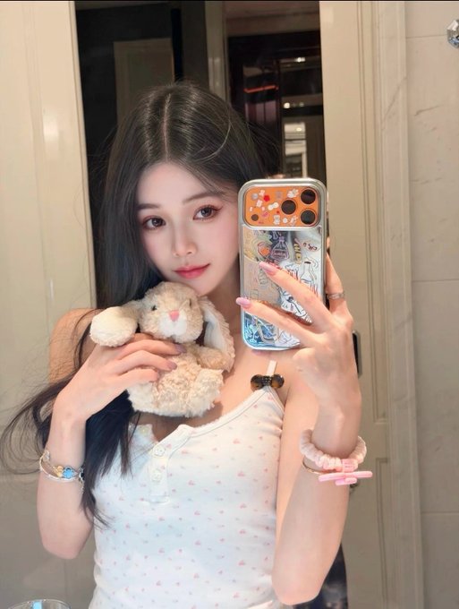 A young woman with long dark hair taking a mirror selfie while holding a stuffed bunny, wearing a white floral top and pink accessories, with an iPhone featuring a colorful case.