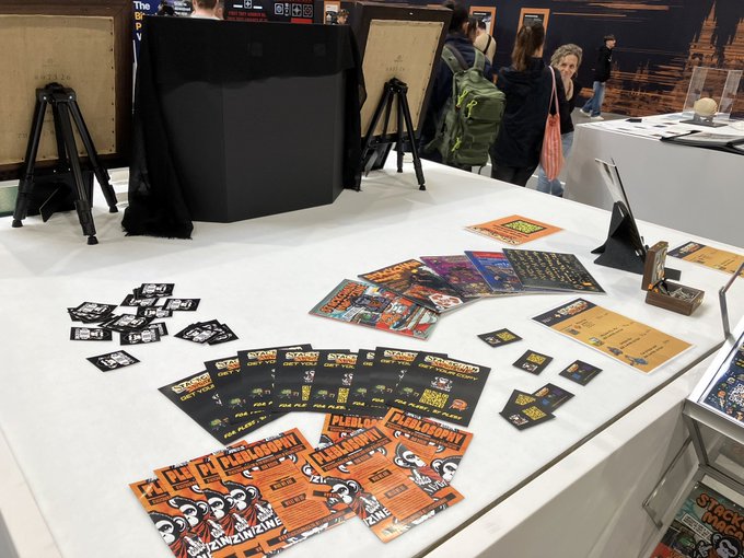 The image shows a table covered with various promotional materials, including posters and flyers for an art exhibition or event. The table has a white surface that contrasts with the colorful and eye-catching designs of the promotional items. There are several people present around the table, possibly discussing the artwork or preparing to attend the event. A backpack can also be seen on the floor near one of the individuals.