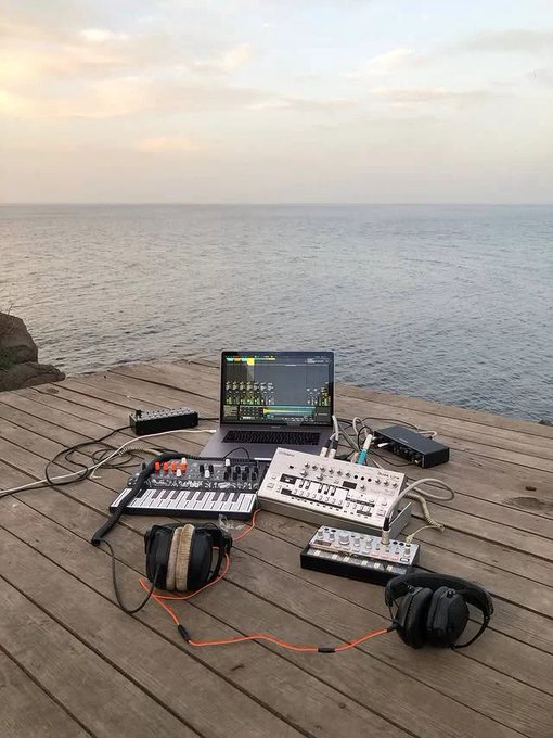 A scenic photo of DJ/music production equipment including a laptop, synthesizers, drum machines, and headphones set up on a wooden deck overlooking the ocean at sunset.