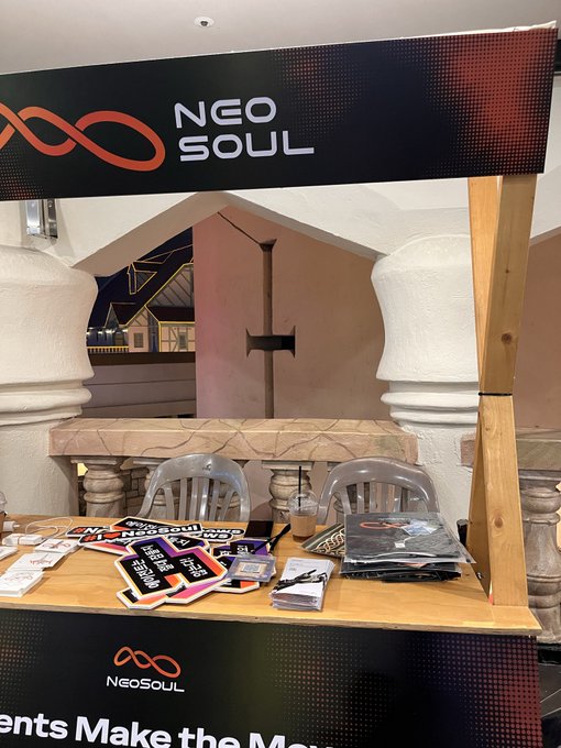 A promotional booth for 'NEOSOUL' set up indoors, featuring a black banner with the company logo, a wooden table with clear chairs, and various branded stickers and merchandise, with partial text reading 'ents Make the Mo...'.