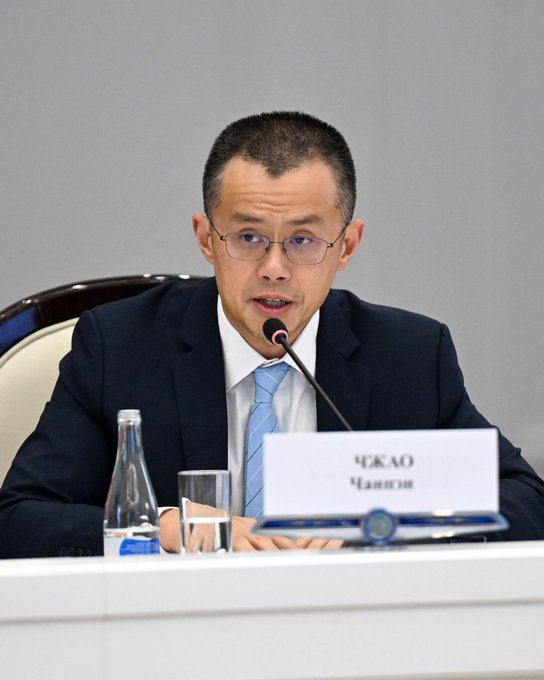 A man with short dark hair, glasses, and a dark suit with a light blue tie sits at a conference table speaking into a microphone, with a name placard reading "ЧЖАО Чанпэн" (Zhao Changpeng) in front of him, alongside a water bottle and glass. The image relates to the accompanying post by identifying the speaker as Changpeng Zhao, the founder of Binance, at a meeting with Kyrgyz President Sadyr Japarov where Binance announced key milestones for Kyrgyzstan, including KGS Token ranking among the top three tokens traded by new users in August, being live on three blockchains, and reaching over 10,000 people through education initiatives.