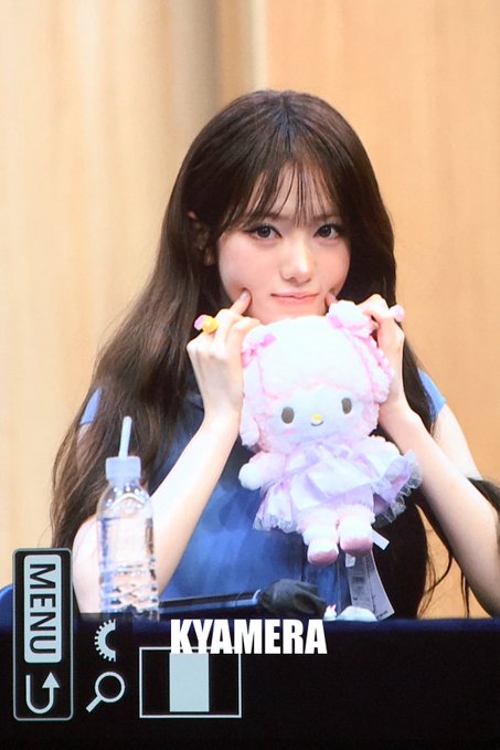 A dark-haired woman holds a pink My Melody plush toy to her face at what appears to be a fan event, with a 'KYAMERA' camera interface overlay visible on the image.