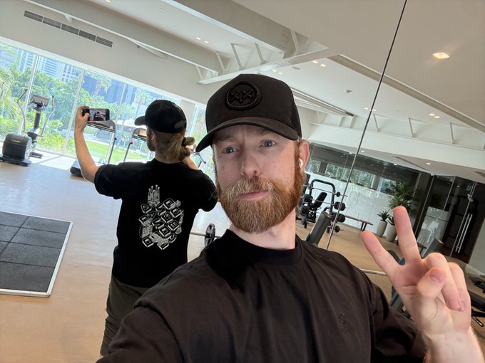 A red-bearded man wearing a black cap and shirt takes a selfie in a gym while flashing a peace sign, with another person in the background taking a photo with their phone. The image visually reinforces the caption "no wimps on @monad" by showcasing the casual, confident gym setting.