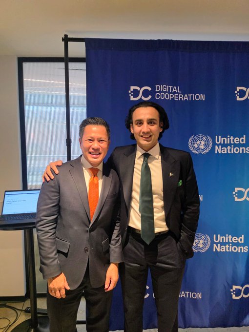 Two men in business suits pose together in front of a 'Digital Cooperation' and 'United Nations' branded backdrop.