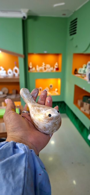 A hand holds a small, carved wooden bird-like figurine in front of a green-walled shop displaying pottery on illuminated shelves.