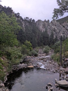 A scenic landscape photograph captures a rocky river flowing through a canyon, surrounded by steep, jagged cliffs and green vegetation under an overcast sky.