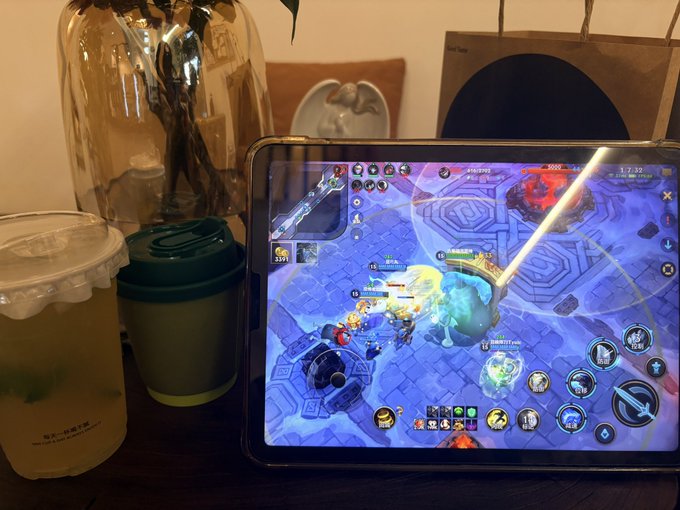 The image shows an open tablet computer displaying a video game called "Crypto Survival Plan" with a player's character standing in front of a blue background, holding a sword. The tablet is placed on top of a wooden table next to two cups filled with drinks and a vase containing flowers. A brown paper bag can also be seen nearby. The post about the new coffee shop suggests that people might gather there for socializing or enjoying some refreshments while playing video games together, creating an inviting atmosphere for gamers to relax and have fun.